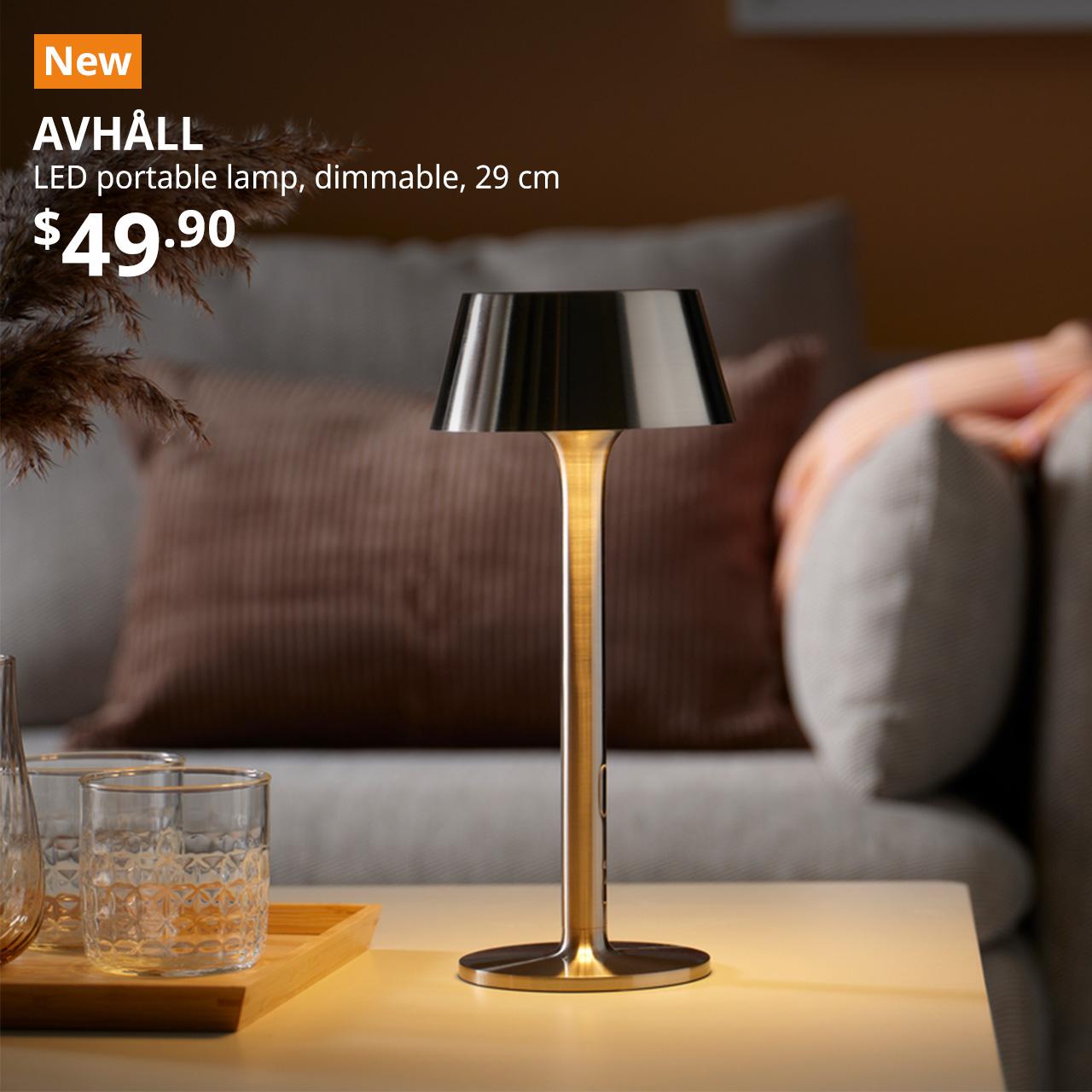 A nickel-plated AVHÅLL LED portable lamp is lit and placed on the coffee table next to a tray of glasses in a living room setting. An orange "NEW" label is displayed in the top left corner of the image, featuring white text that reads: "AVHÅLL, LED portable lamp, battery-operated dimmable, 29 cm, $49.90"