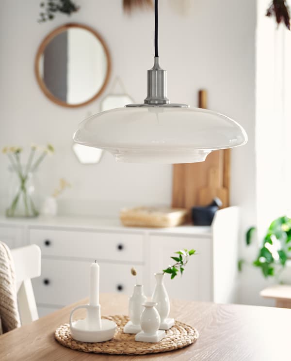 A nickel-plated pendant lamp with a white lampshade in glass hangs over a table top in oak veneer where white vases stand.