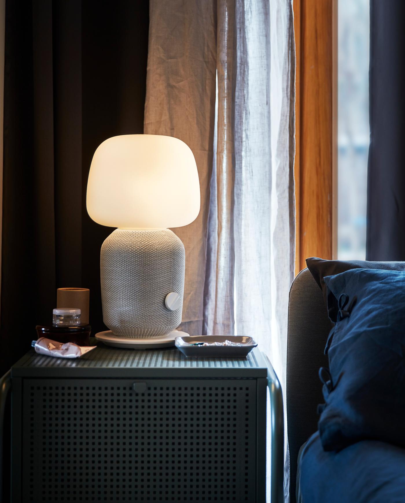 A night stand with a white and grey SYMFONISK table lamp with WiFi speaker standing in front of a window with grey curtains.