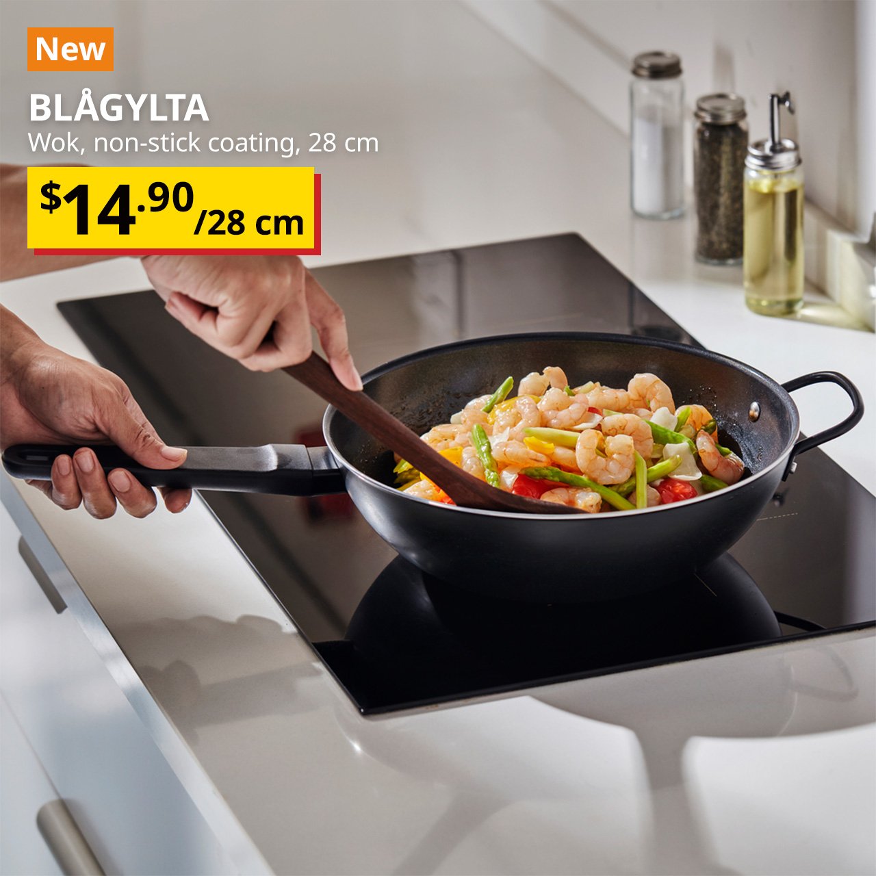 A non-stick coating black BLÅGYLTA wok on a induction top while prawn and asparagus are being stir-fried. Text on the top left of the image reads: BLÅGYLTA, Wok, non-stick coating, 28 cm. Price: $14.90/28 cm.