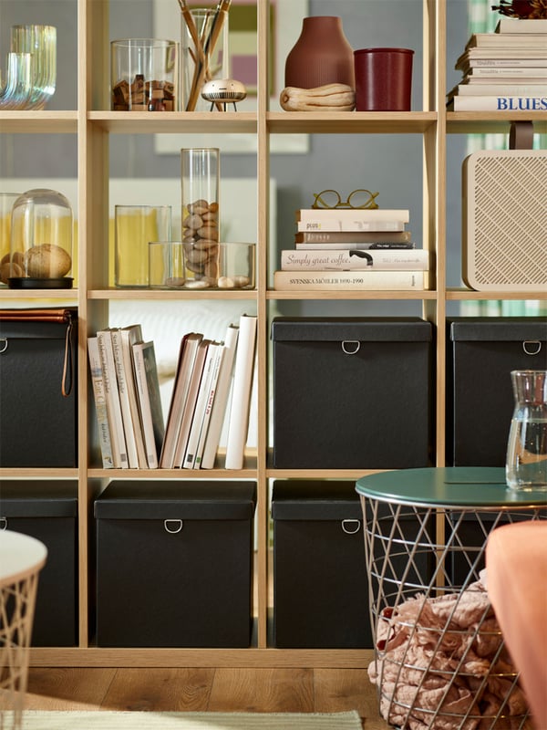 A oak-coloured KALLAX shelving unit with 4-by-4 square compartments. Each compartment is filled with stacks of books, black boxes, or vases and other decorations.