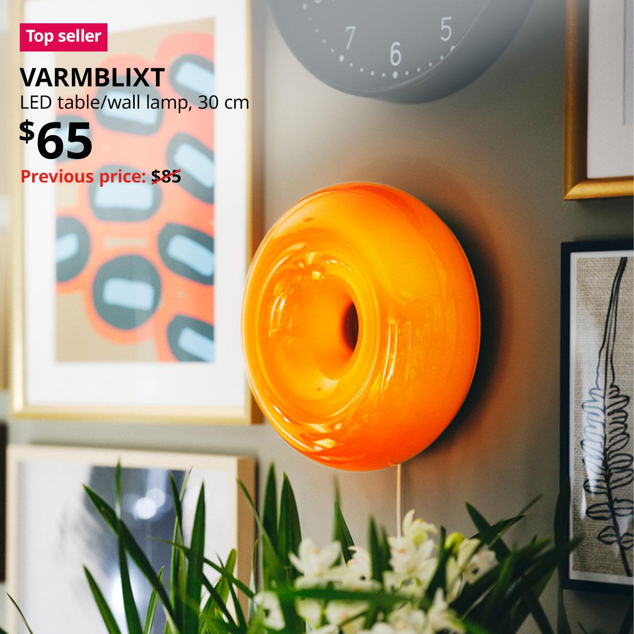 A orange glass VARMBLIXT LED table/wall lamp hung on the wall with photo frames display on the wall. A red "Top seller" label is on the top left of image, with black text below that reads: VARMBLIXT, LED table/wall lamp, 30 cm. Price at $65, with previous price slashed $85.