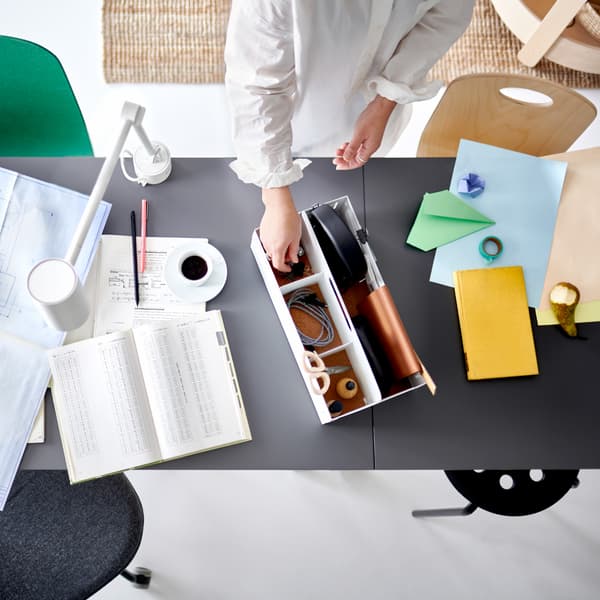A pair of hands picking an item from a white desk organiser on two joined table tops with work papers and a white work lamp.