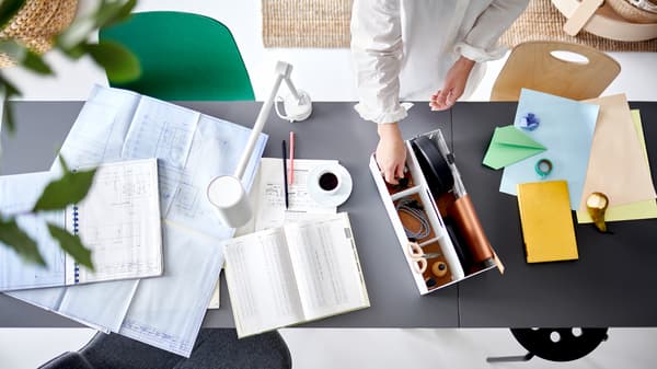 A pair of hands picking an item from a white desk organizer on two joined table tops with work papers and a white work lamp.