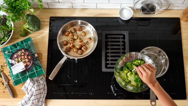 A pan is cooking HUVUDROLL plant balls in sauce on an induction hob. Beside is a strainer with broccoli.