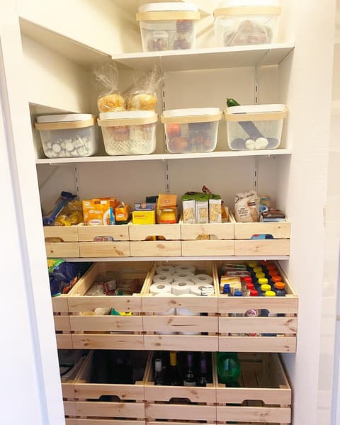 A pantry that is neatly organised with KNAGGLIG wooden crates and RISATORP baskets containing household supplies, such as toilet rolls and soft drinks.