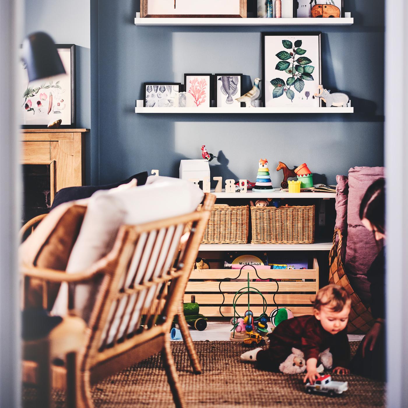 A parent and child in a living room with white MOSSLANDA picture ledges, frames, rattan baskets, armchairs, a rug and toys.
