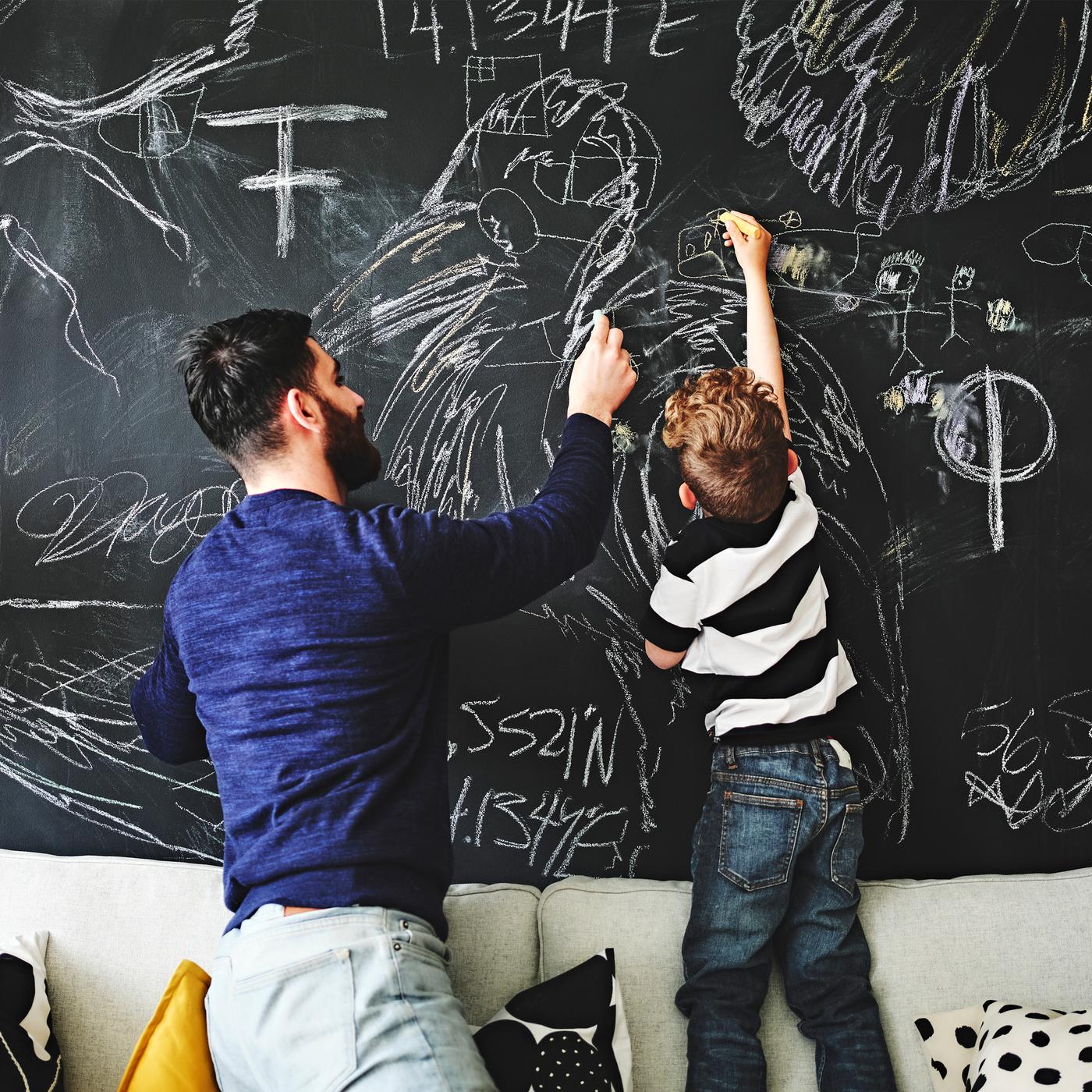 A parent and child kneel and stand together on top of a sofa, drawing on a wall covered in blackboard paint.