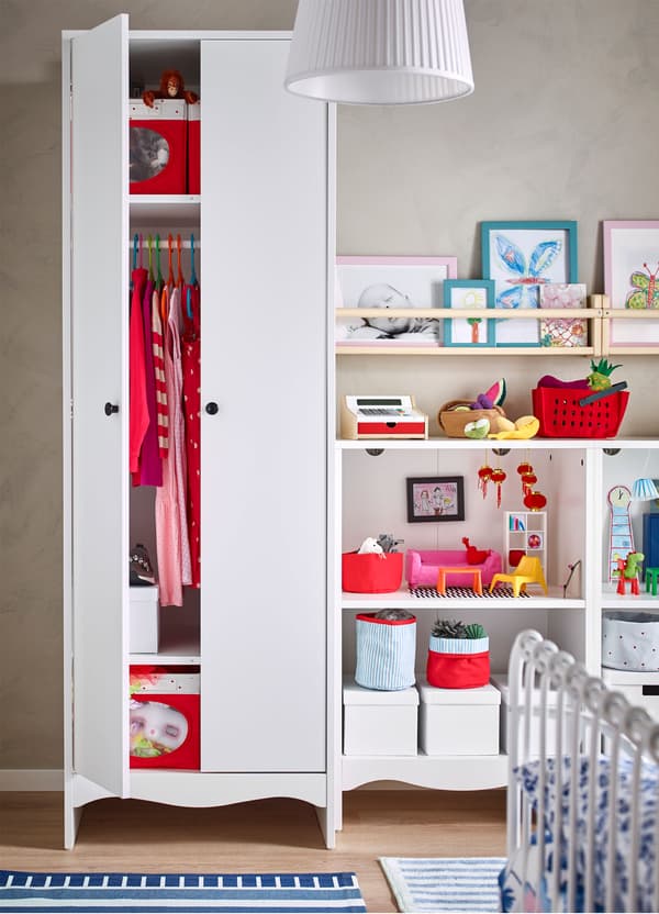 A partially opened IKEA SOLGUL white children’s wardrobe, revealing clothes and storage boxes, beside open storage with toys.