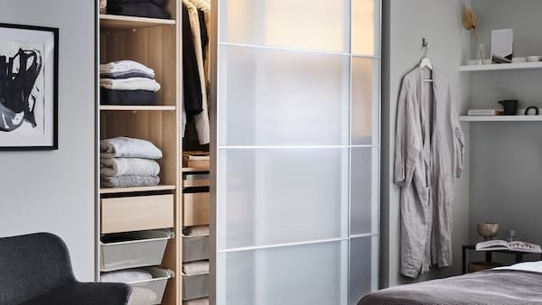 A partly open PAX/SVARTISDAL wardrobe with shelves, drawers and hanging clothes stands in a bedroom near a bed.