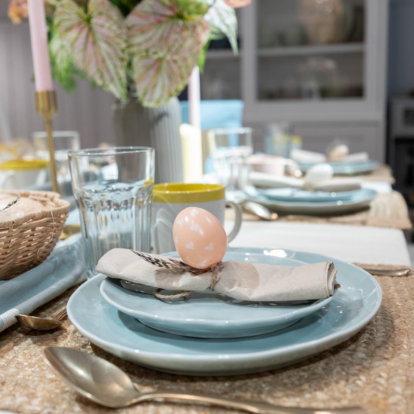 A pastel blue place setting with a napkin topped by a decorative Easter egg, set on a woven placemat with glassware and candles in the background.
