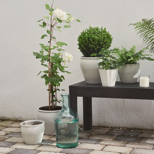 A patio area features a white AMBRARÖNN plant pot, surrounded by lush green plants and outdoor decor.