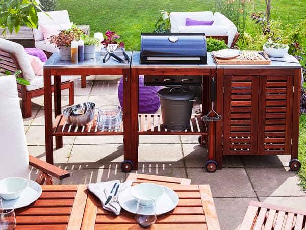 A patio with an ÄPPLARÖ/KLASEN charcoal barbecue with trolley and cabinet in acacia, and in front is an outdoor dining table.