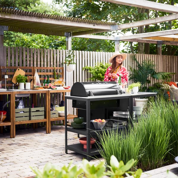 A patio with greenery showcases a black GRILLSKÄR charcoal barbecue on a kitchen island, a woman standing next to it.