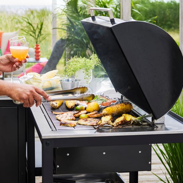 A patio with lush greenery features a black GRILLSKÄR charcoal barbecue made of black and stainless steel cooking food.