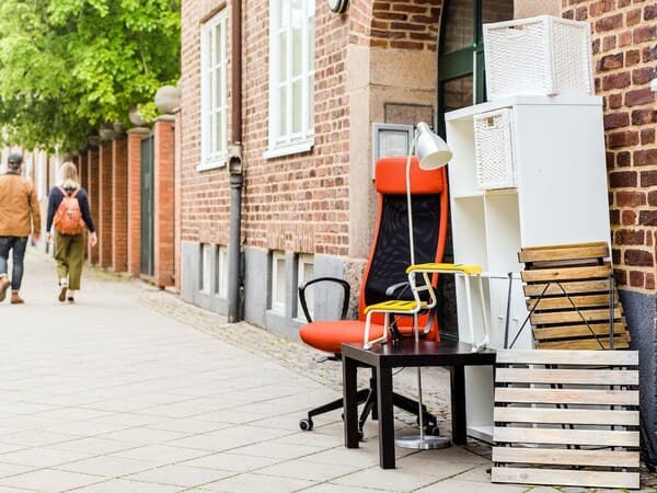 A pavement next to a brick building with furniture left out: an orange and black office chair, a small table with a white lamp, a shelf with baskets and wooden slats; two passers-by in the background.