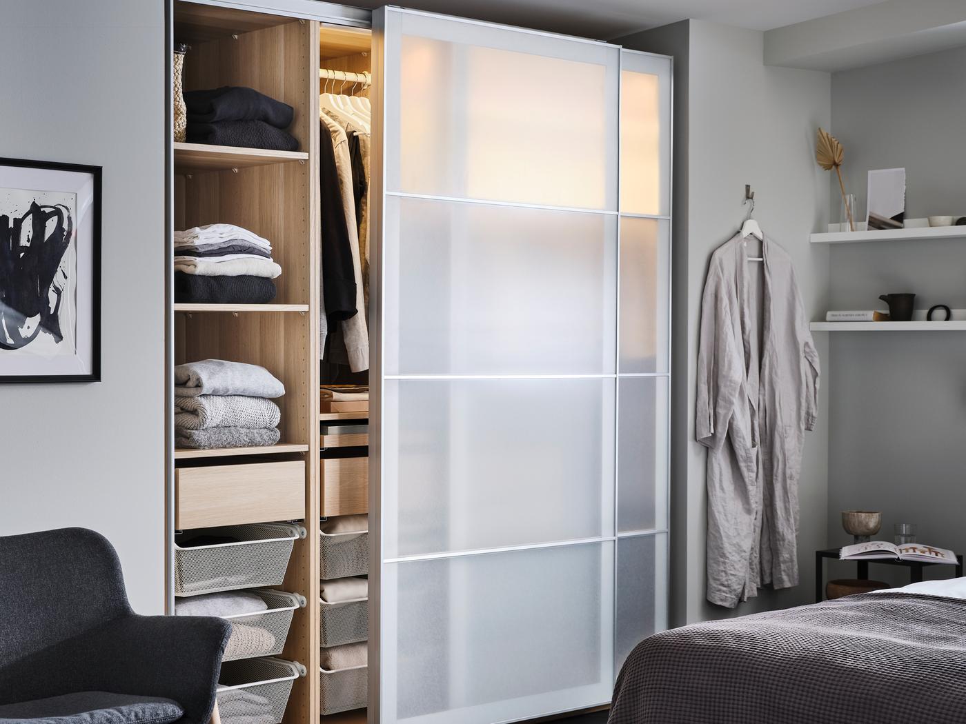 A PAX/SVARTISDAL wardrobe in a grey bedroom with one door open showing clothes in piles on shelves and on a hanging rail.
