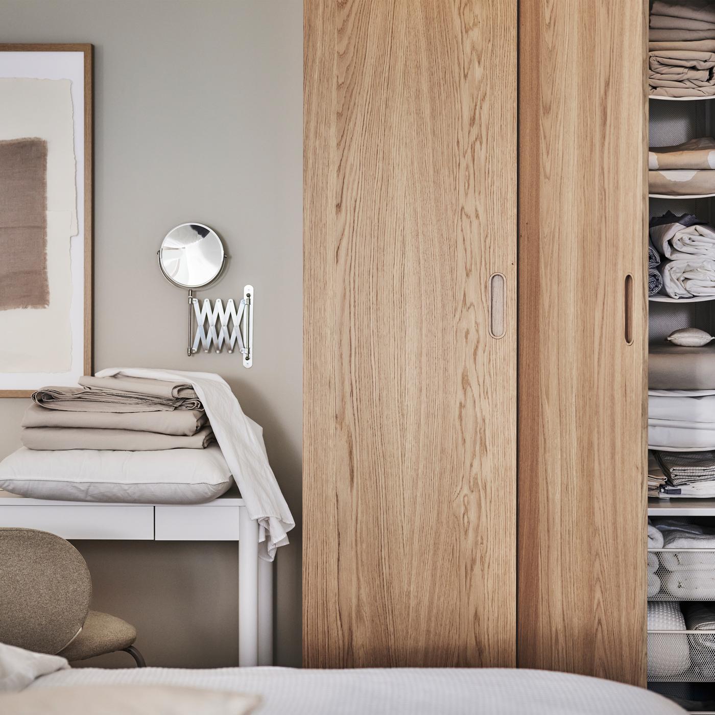 A PAX/TONSTAD wardrobe in white/oak veneer in front of a bed with one door open showing shelves holding folded textiles.