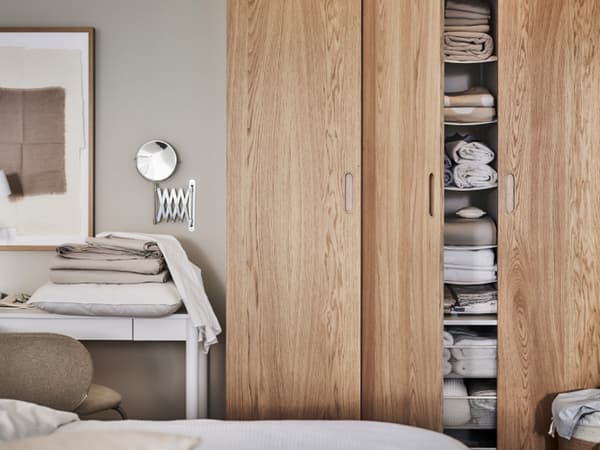 A PAX/TONSTAD wardrobe in white/oak veneer with one door open showing shelves holding folded textiles, in front of a bed.