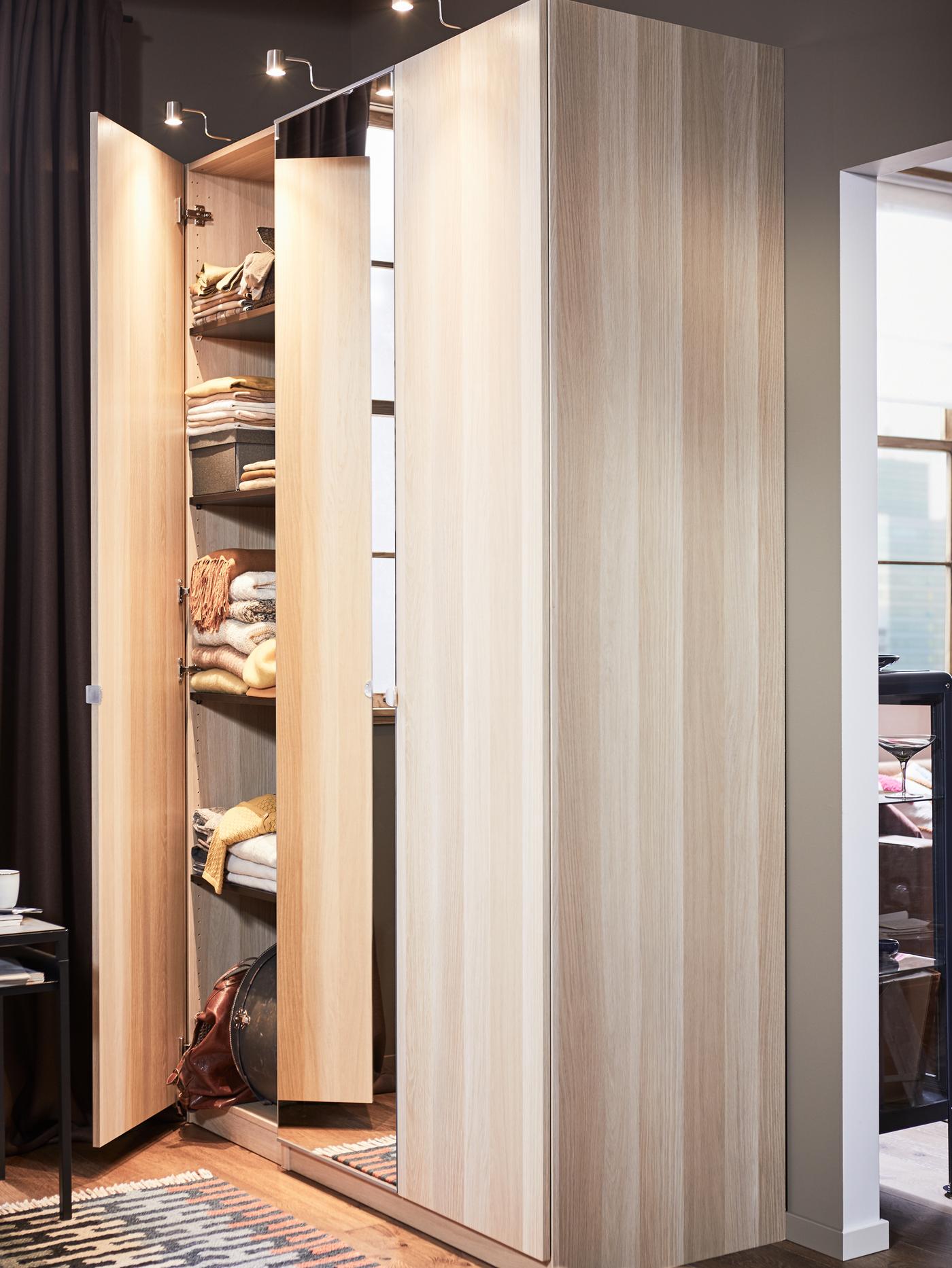 A PAX wardrobe in white stained oak with one door ajar. Inside the wardrobe are several shelves of clothes.