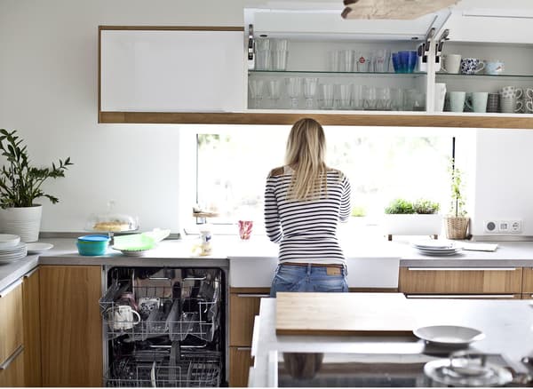 A person at the sink in a predominantly white kitchen that has wood cabinet fronts.