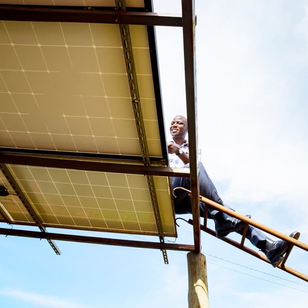 A person atop a ladder inspects the top side of a solar panel on a bright sunny day.