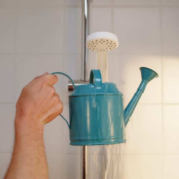 A person fills a teal watering can under a shower head in a tiled bathroom.