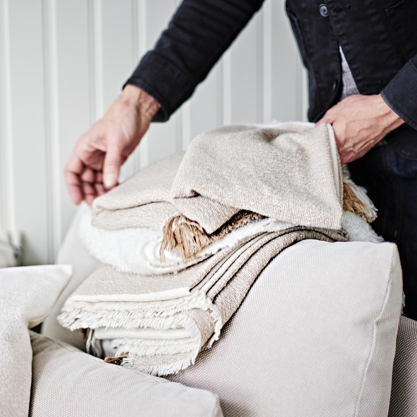 A person folding a grey-beige GUNNEMOR throw on the arm of a light beige SÖDERHAMN 1-seat corner section.