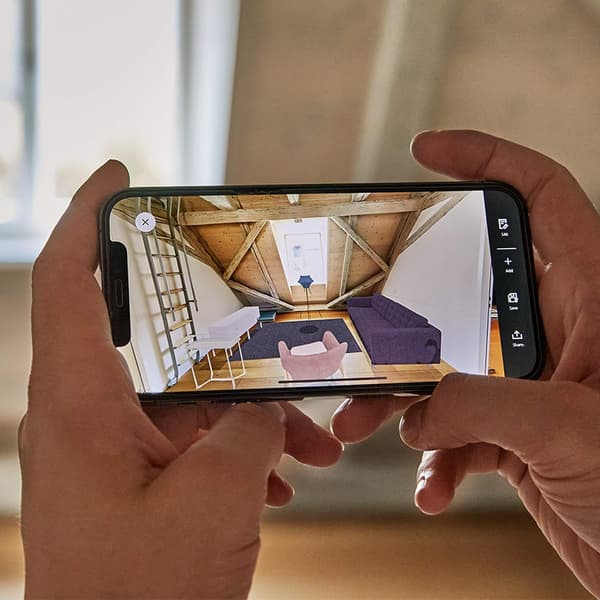 A person holding a smartphone which shows an empty room furnished with IKEA products in augmented reality.