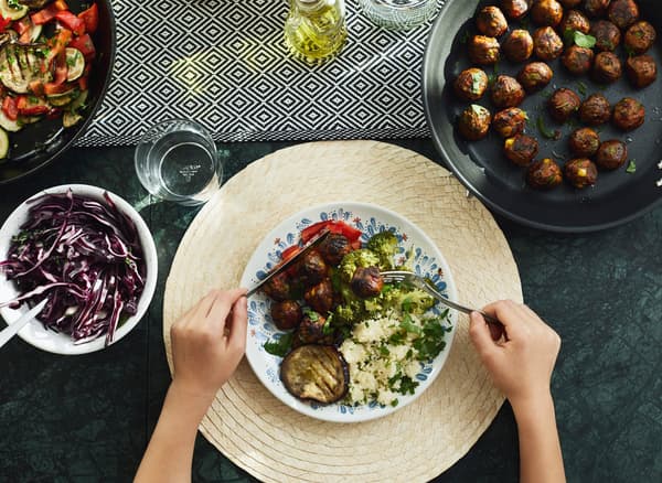 A person holds a knife and fork over a plate of food on a table with serving dishes of vegetables and veggie balls.