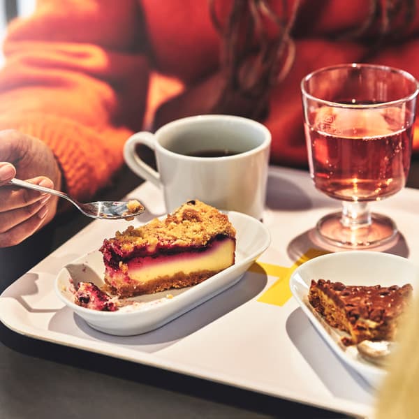 A person holds a spoon over a slice of berry cheesecake on a Swedish Restaurant table with coffee, soft drinks and another slice of cake nearby.