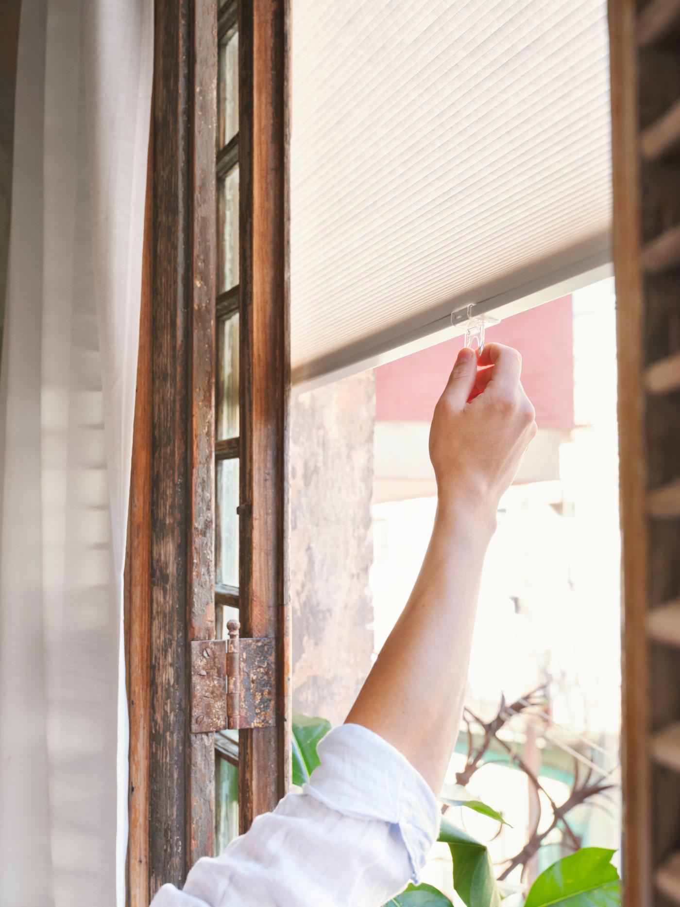 A person holds the tab of a white HOPPVALS cellular blind that hangs at a window with an aged-looking wooden frame.
