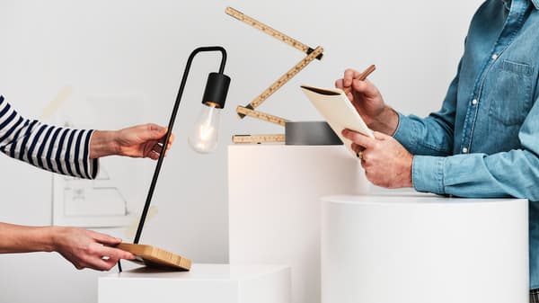 A person in a blue denim shirt writes in a notebook as another person holds out a black/bamboo TVÄRHAND table lamp.
