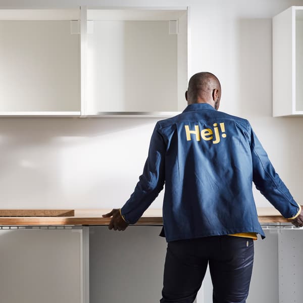 A person in a blue jacket installing a white kitchen with base and top cabinets and an oak effect/laminate worktop.