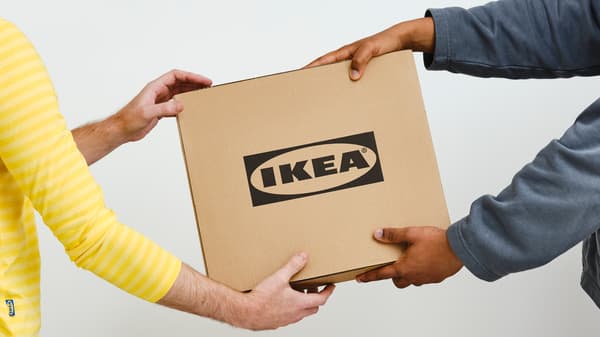 A person in a gray sweater hands a square cardboard box with a black IKEA logo to a person in a yellow IKEA co-worker top.