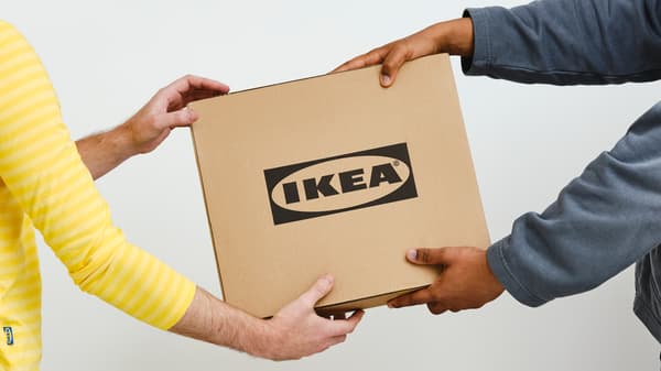 A person in a gray sweater hands a square cardboard box with a black IKEA logo to a person in a yellow IKEA co-worker top.