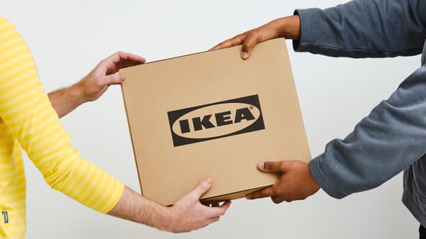 A person in a gray sweater hands a square cardboard box with a black IKEA logo to a person in a yellow IKEA co-worker top.