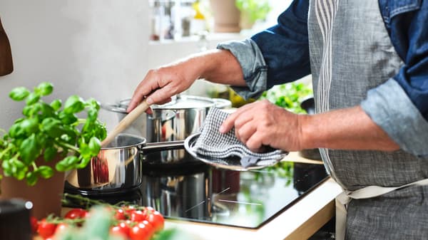 A person in a grey MARIATHERES apron stirs with a wooden spoon a saucepan at the back of a black MATMÄSSIG induction hob.