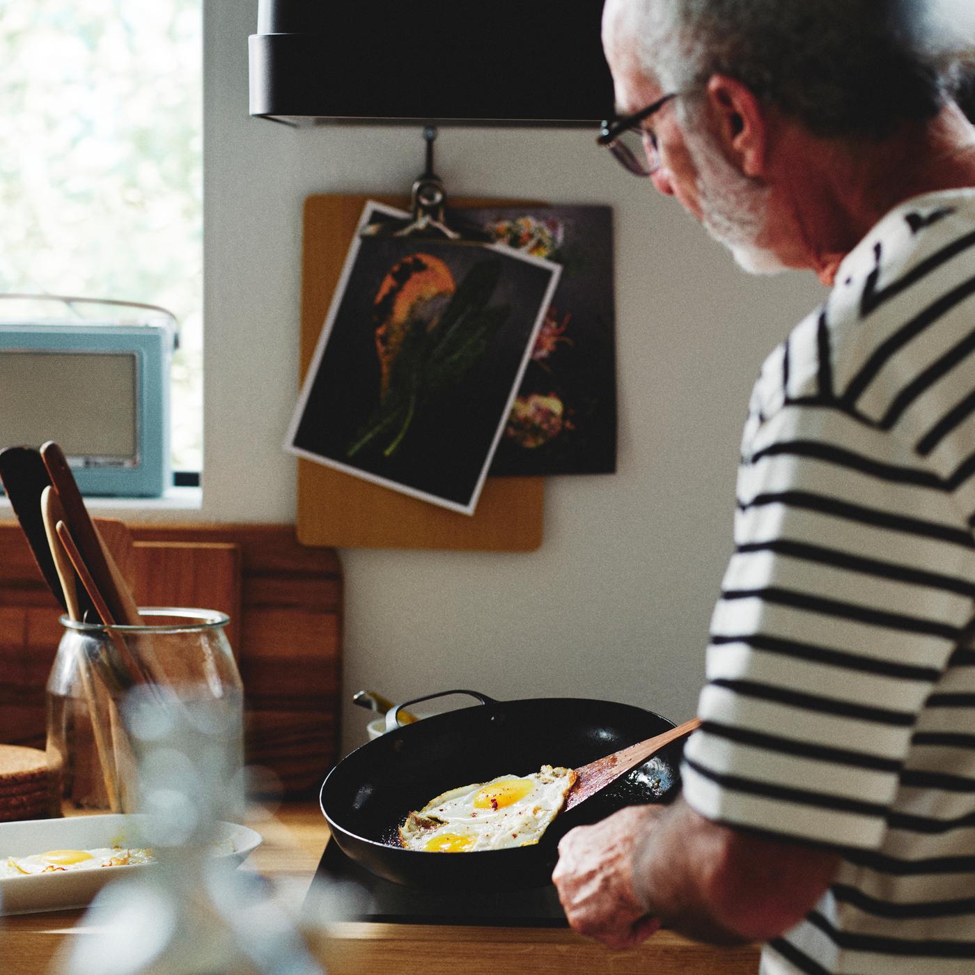 A person in a kitchen frying an egg in a carbon steel VARDAGEN frying pan next to a VARDAGEN jar with lid holding utensils.