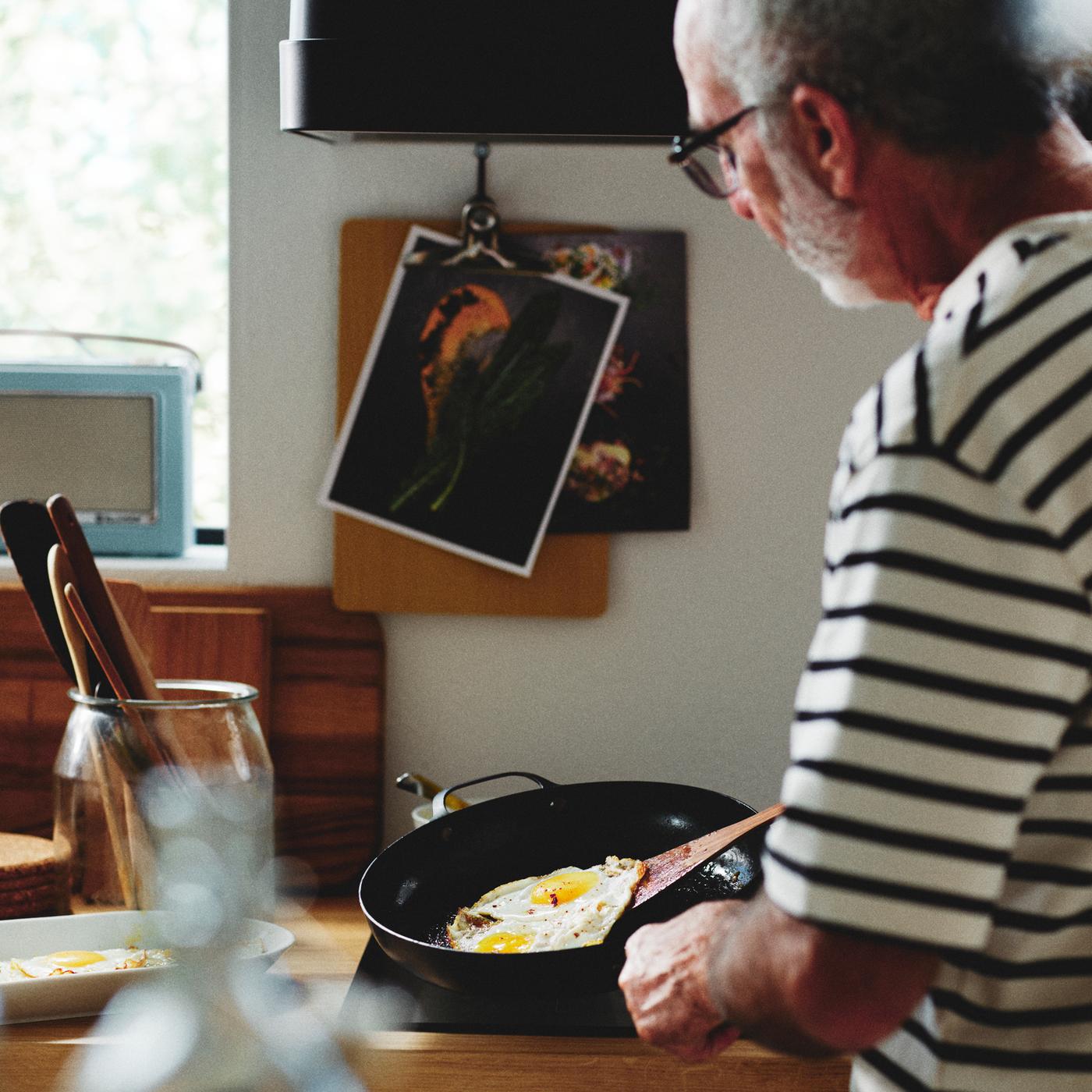 A person in a kitchen frying an egg in a carbon steel VARDAGEN frying pan next to a VARDAGEN jar with lid holding utensils.
