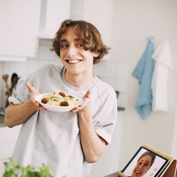 A person in a kitchen holds up a plate of food while video calling someone on a tablet that’s propped up on the counter. Fresh herbs, jars, and cooking ingredients are spread out on the workspace.