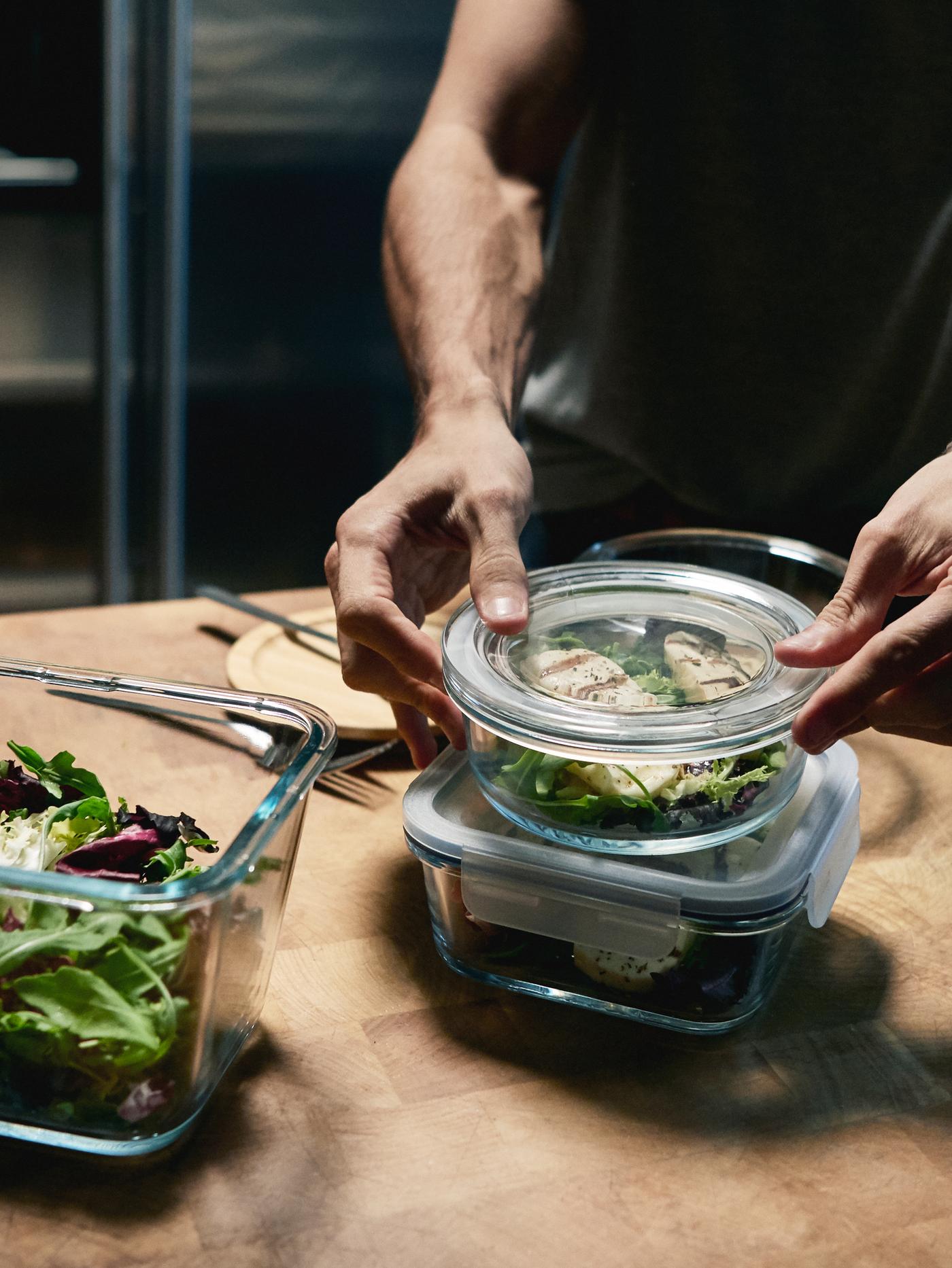 A person in a kitchen putting the lid on a glass container of food. On the same benchtop are two other full food containers.