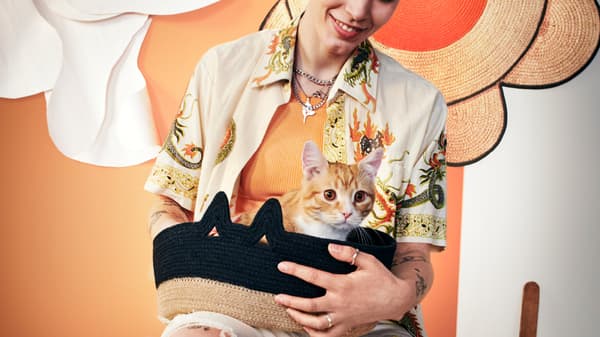 A person in a patterned short-sleeved shirt is holding a MÄVINN basket black/natural with an orange and white cat in it.