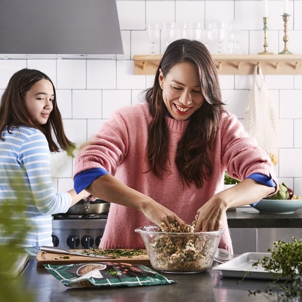 A person in a pink sweater prepares food in a kitchen, mixing ingredients in a bowl on the counter, while another person stands nearby at the stove.