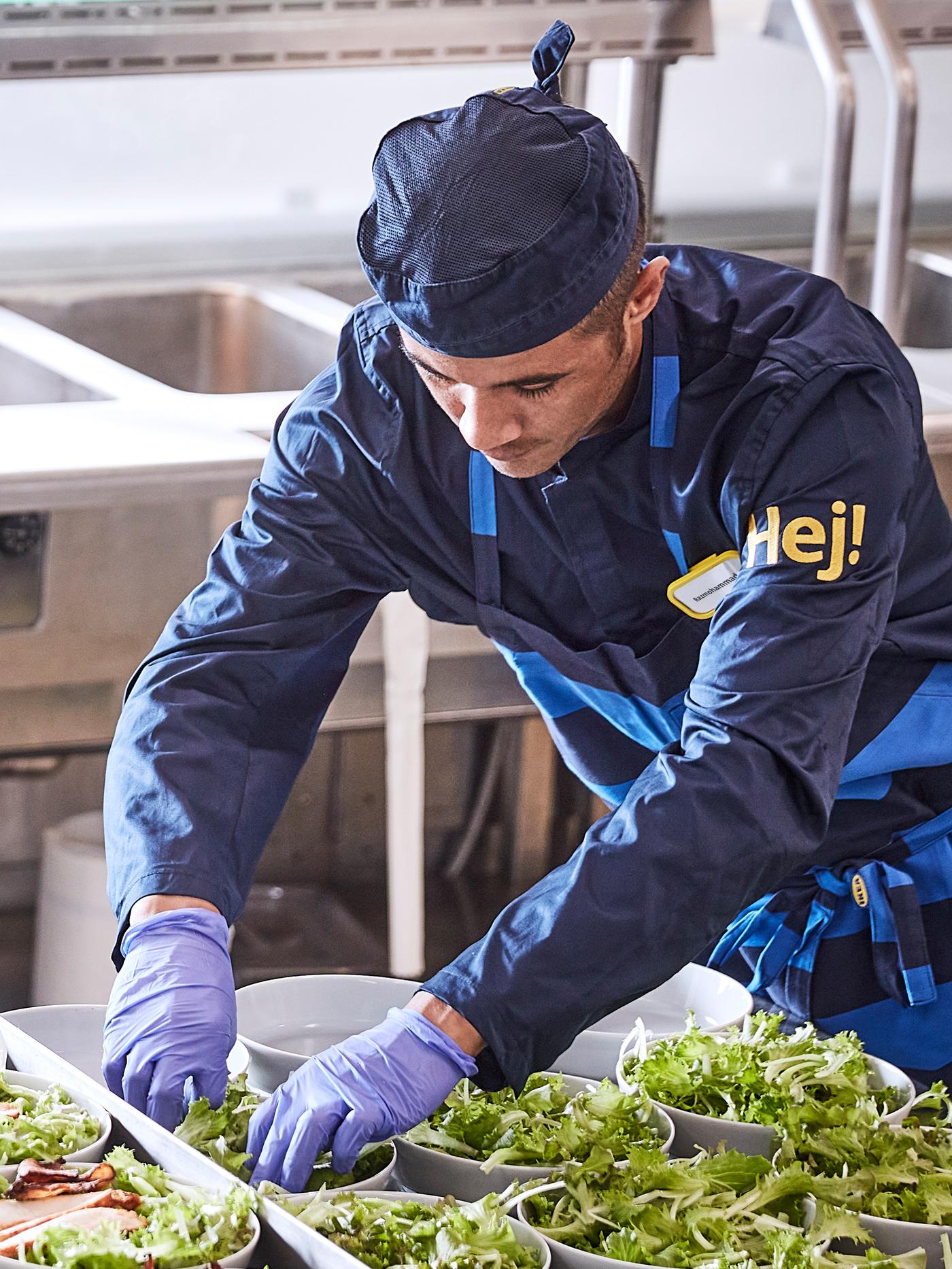 A person in a restaurant kitchen wearing work clothes, gloves and an apron is filling bowls in a row with salad.