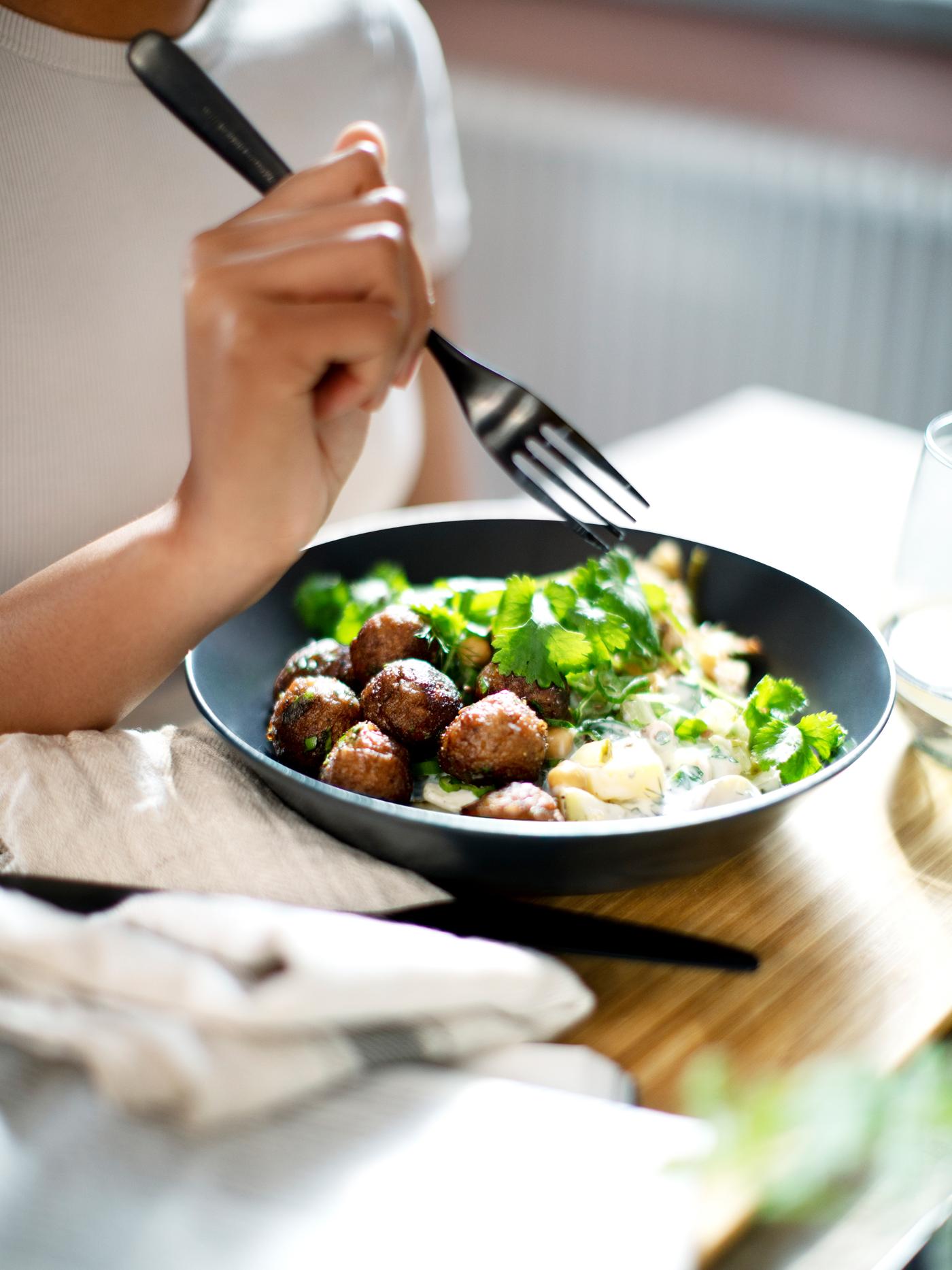 A person in a white t-shirt holds a fork over a dish of HUVUDROLL plant balls, potatoes, sauce and fresh herbs.