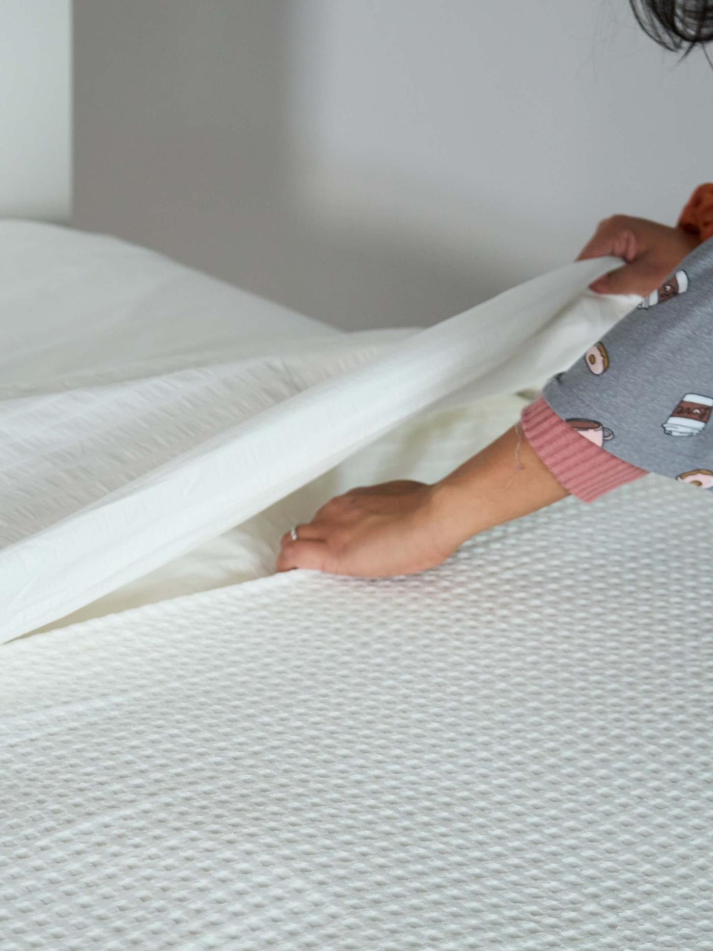 A person in pajamas making a bed, lifting a white bedsheet over a textured mattress cover in a clean, minimal bedroom setting.