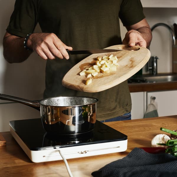 A person is dropping chopped potatoes from a chopping board into a chrome casserole on a TILLREDA portable induction hob.