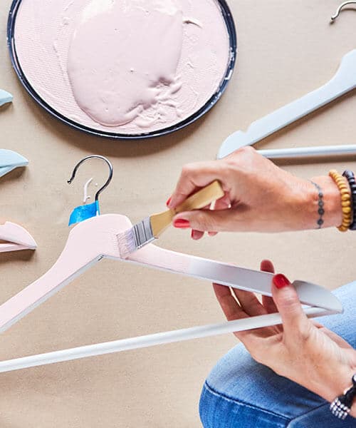 A person is holding a white BUMERANG hanger and is painting it light pink. There are other hangers nearby, some painted light pink and some painted light blue.