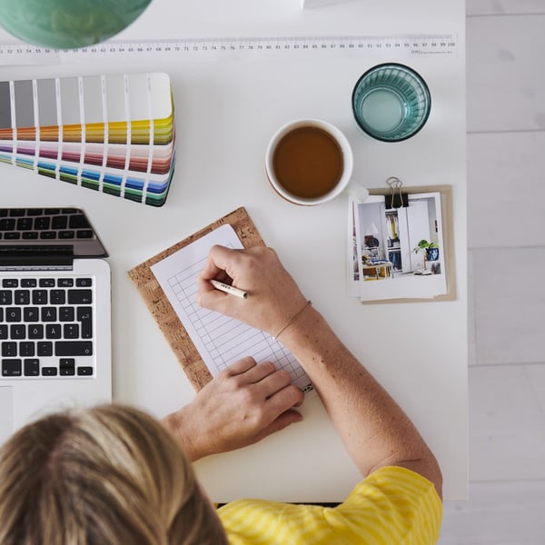 A person is writing on a note book next to a cup of tea and color palet